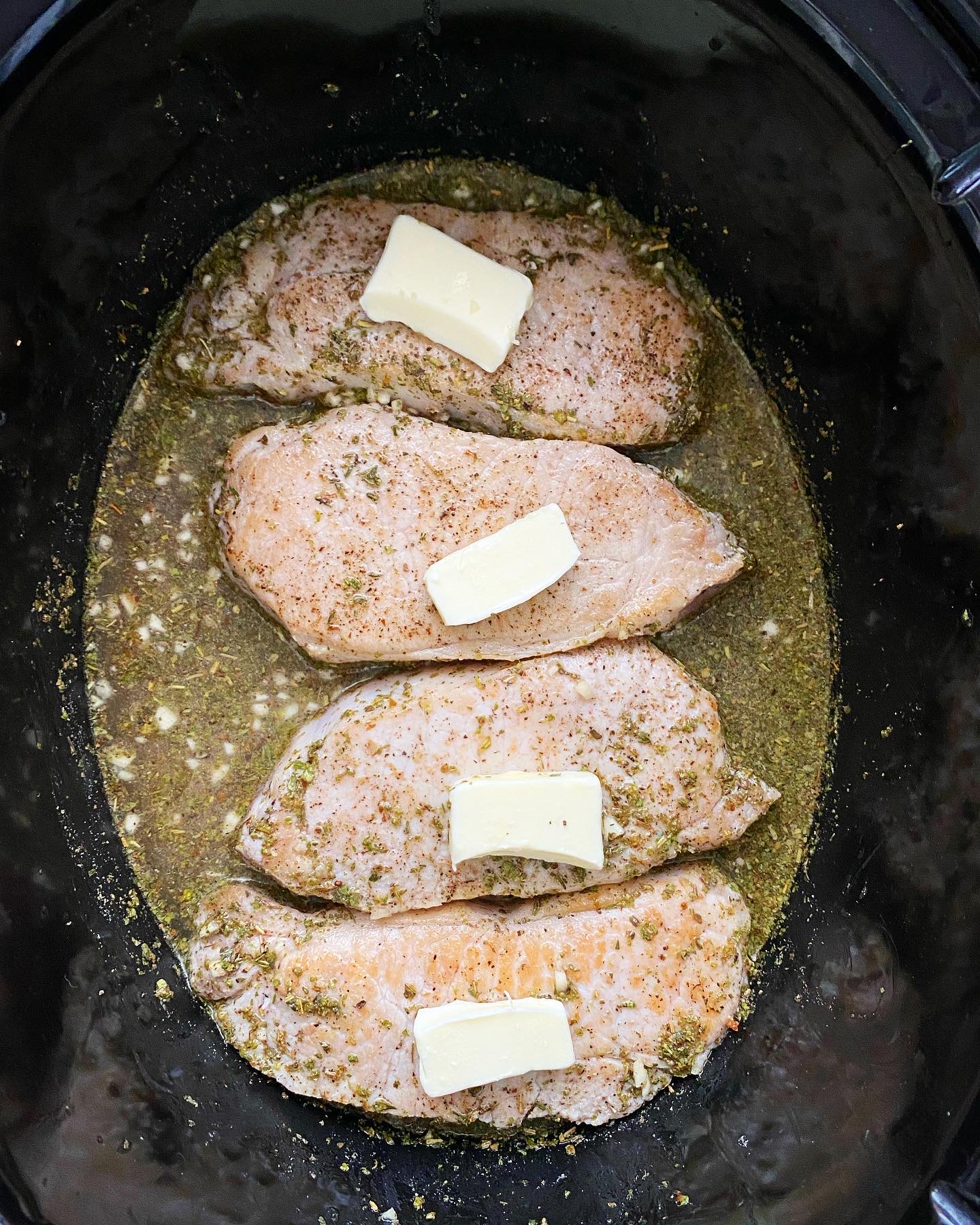 overhead shot of uncooked, seared pork chops in the a black slow cooker with butter on top