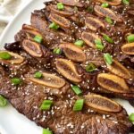 close up shot of cooked kalbi short ribs on a white plate garnished with sesame seeds and green onions