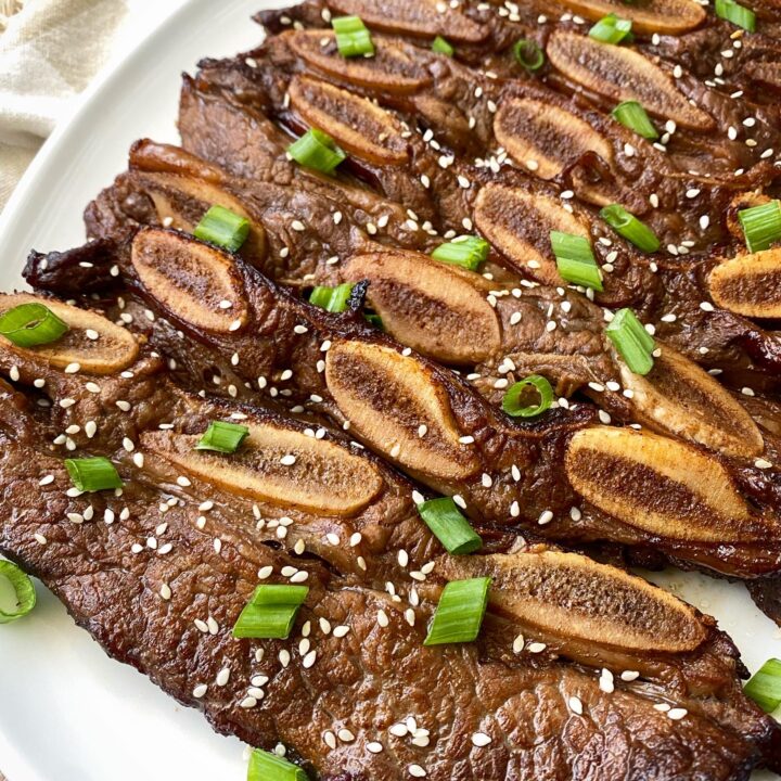 close up shot of cooked kalbi short ribs on a white plate garnished with sesame seeds and green onions