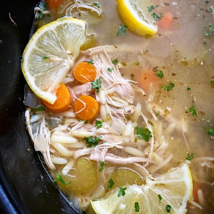 overhead shot of lemon Chicken Orzo Soup in a black slow cooker garnished with a lemon slice