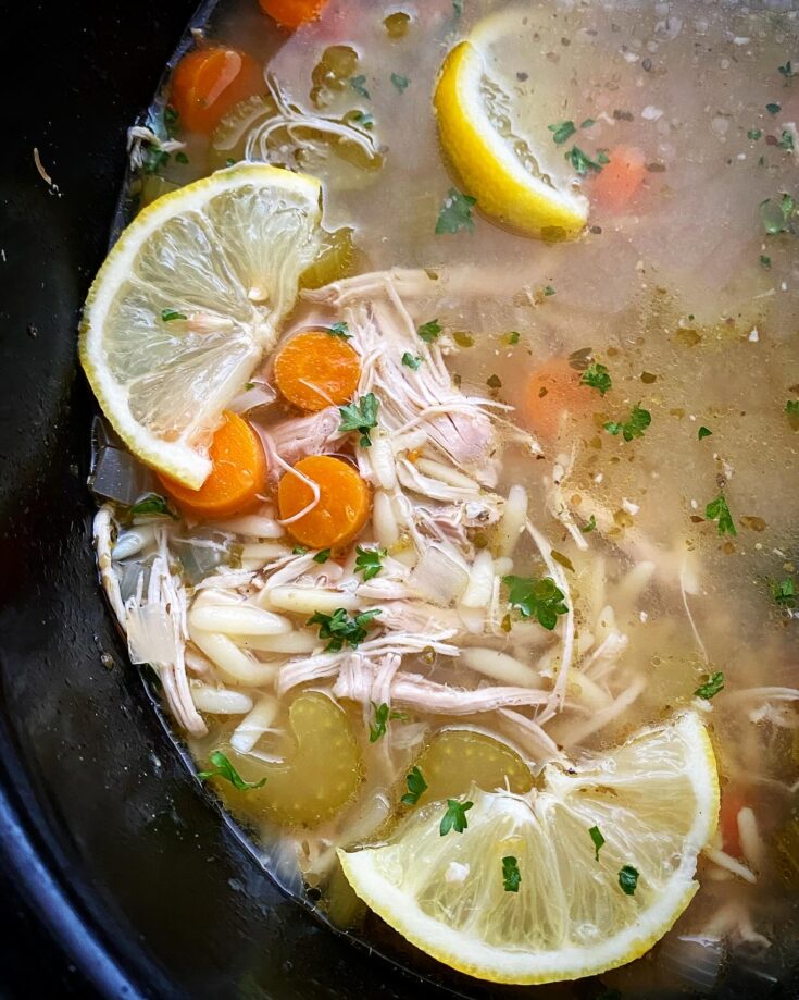overhead shot of lemon Chicken Orzo Soup in a black slow cooker garnished with a lemon slice