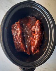 overhead shot of uncooked beef back ribs in a black slow cooker with BBQ sauce