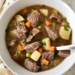overhead shot of cooked steak & potato soup in a white bowl with a silver spoon