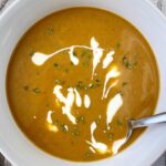 overhead shot of pumpkin bisque in a white bowl garnished with fresh thyme and a drizzle of coconut milk