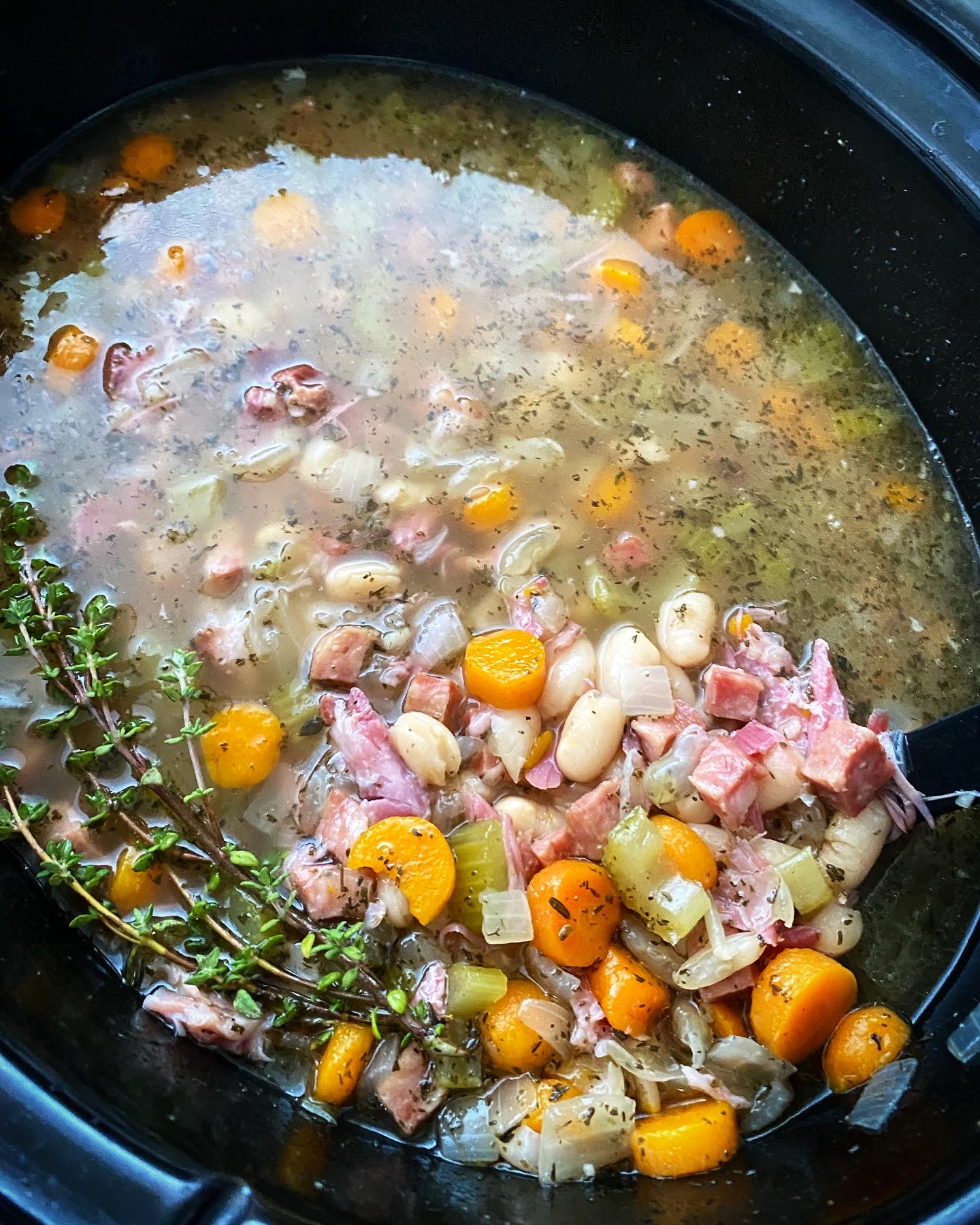 close up shot of cooked crockpot ham and bean soup in a black slow cooker garnished with fresh thyme