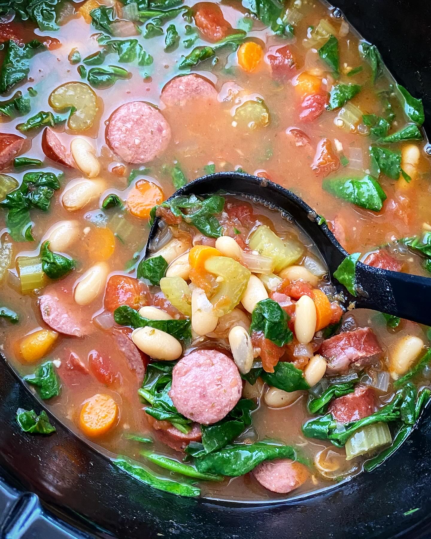overhead shot of cooked Crockpot Sausage Bean Veggie Soup in a black slow cooker with fresh spinach