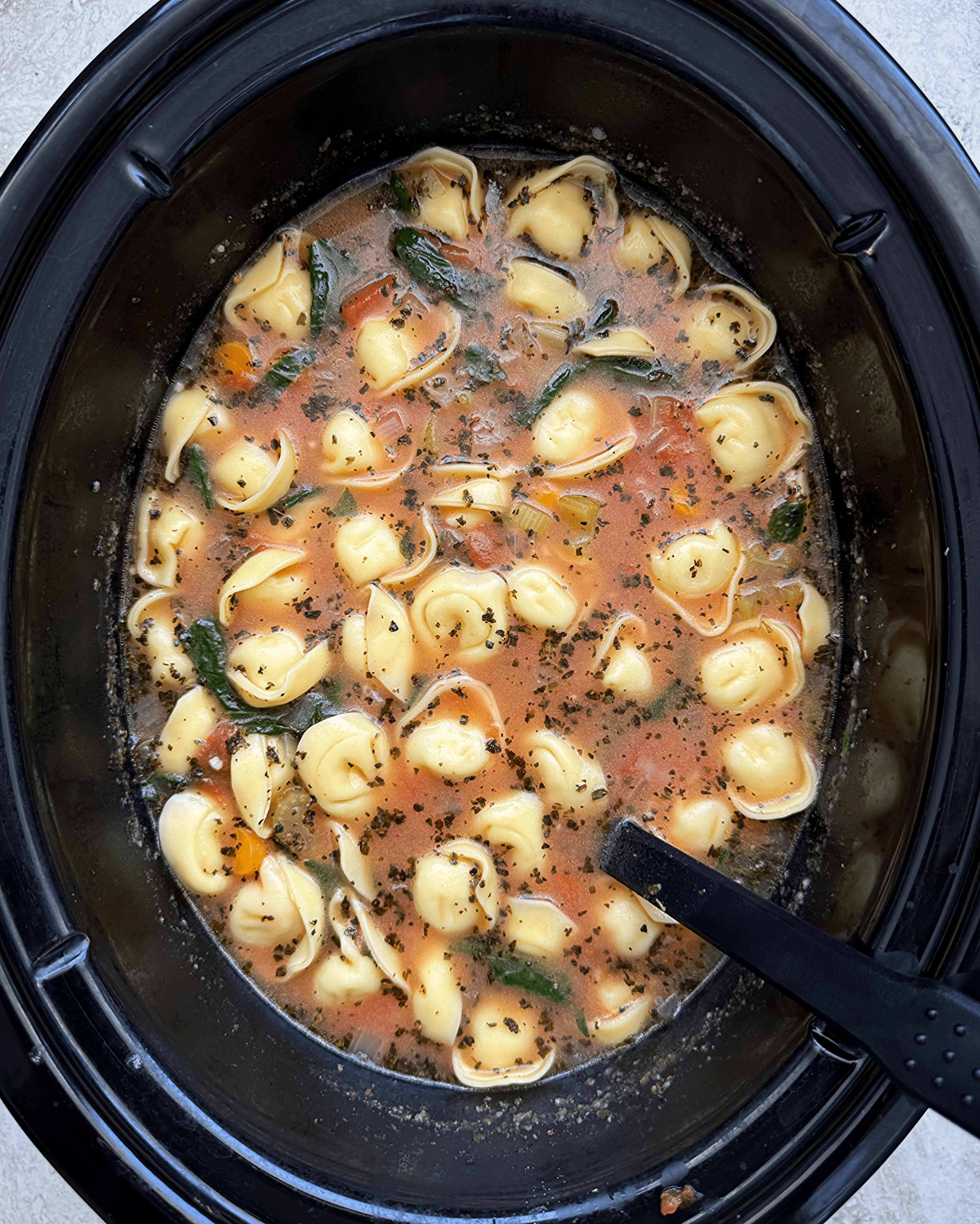 overhead shot of vegetable tortellini soup in a black slow cooker with a black spoon