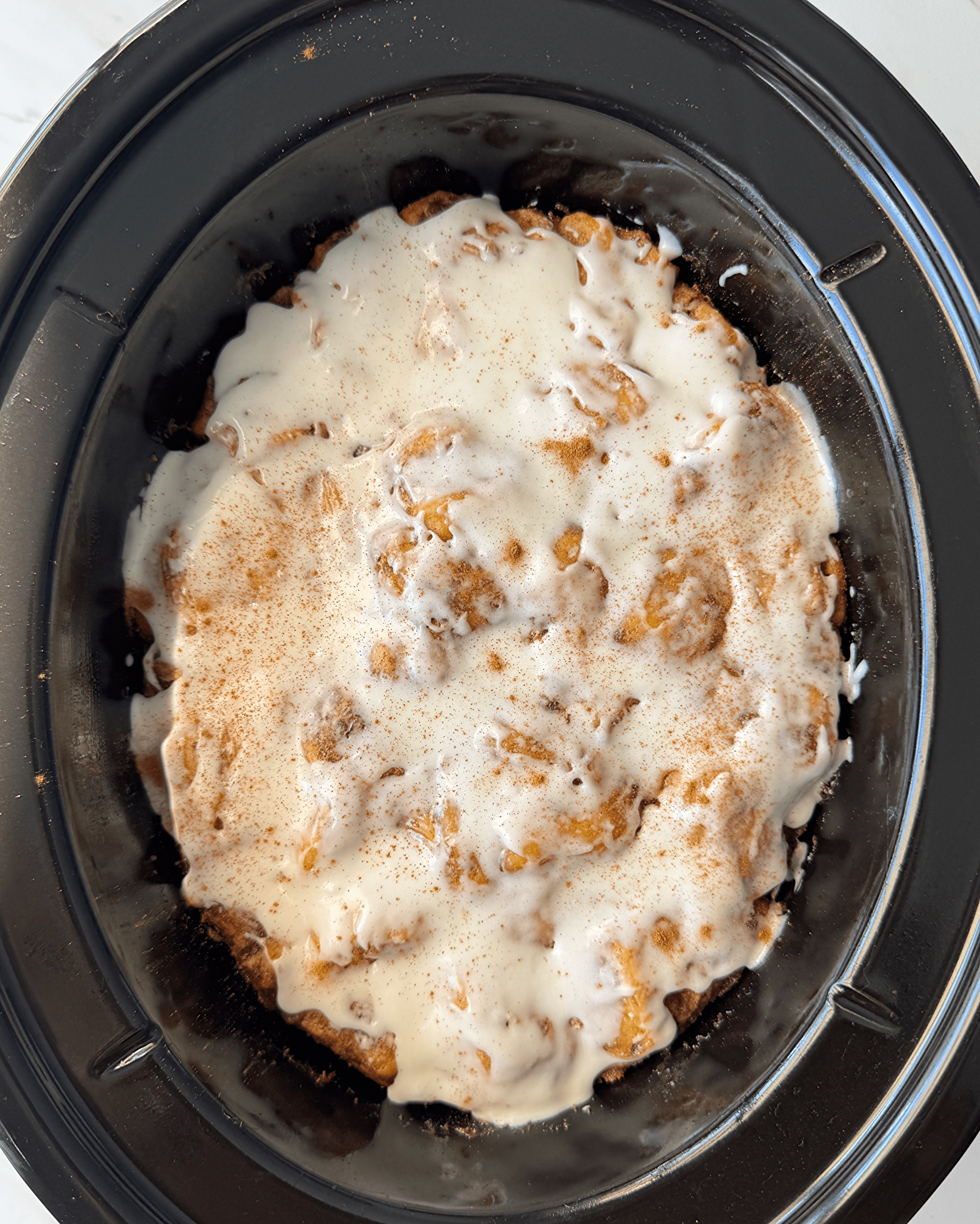 overhead shot of cooked Cinnamon Roll Casserole in a black slow cooker with icing and cinnamon sprinkled on top
