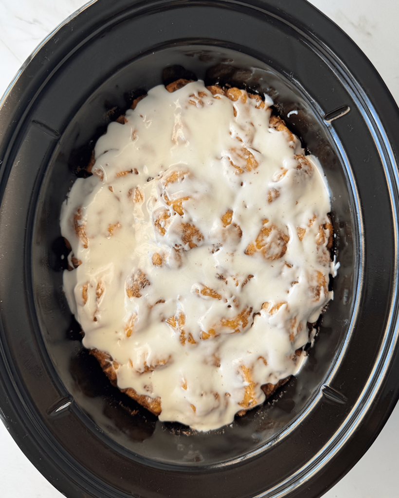 overhead shot of cooked Cinnamon Roll Casserole in a black slow cooker with icing