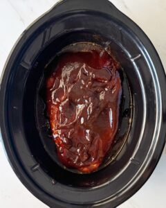 overhead shot of uncooked Beef in a black slow cooker with BBQ sauce on top