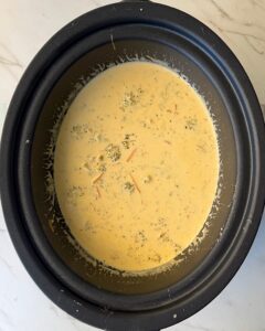 overhead shot of cooked Broccoli Cheddar Soup in a black slow cooker with cream and cheese added