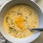 overhead shot of Broccoli Cheddar Soup in a white bowl garnished with cheddar cheese with a silver spoon