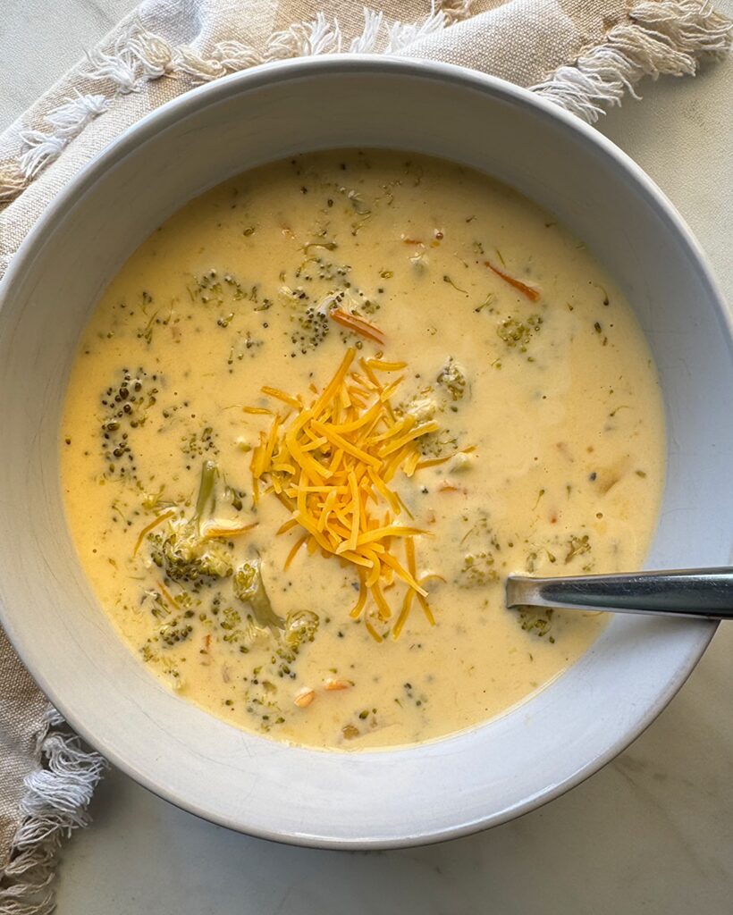 overhead shot of Broccoli Cheddar Soup in a white bowl garnished with cheddar cheese with a silver spoon