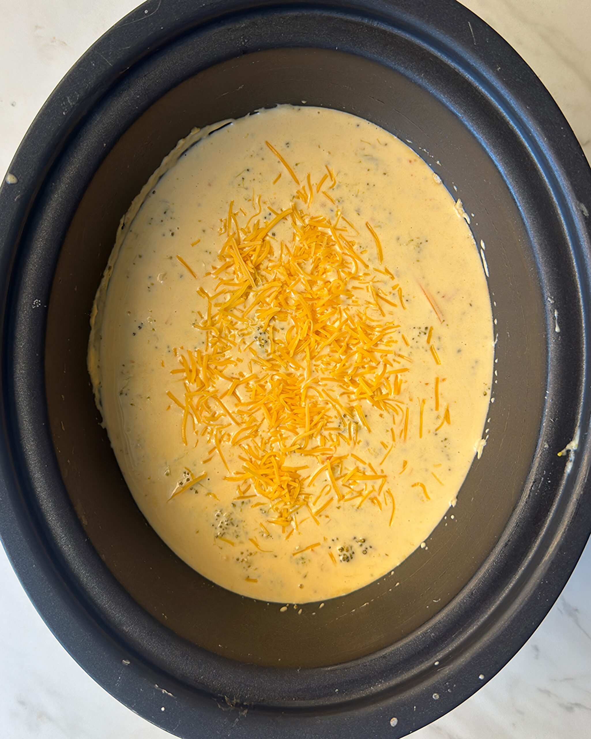 overhead shot of cooked Broccoli Cheddar Soup in a black slow cooker with garnished with shredded cheddar cheese