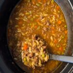overhead shot of cooked Beef Pumpkin Orzo Soup in a black slow cooker with a black spoon