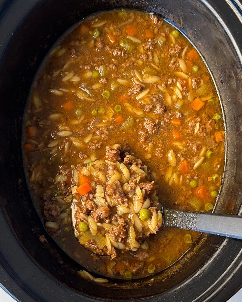 overhead shot of cooked Beef Pumpkin Orzo Soup in a black slow cooker with a black spoon