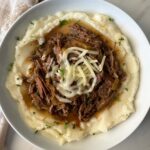 overhead shot of cooked French onion Pot Roast over mashed potatoes in a white bowl garnished with melted shredded cheese