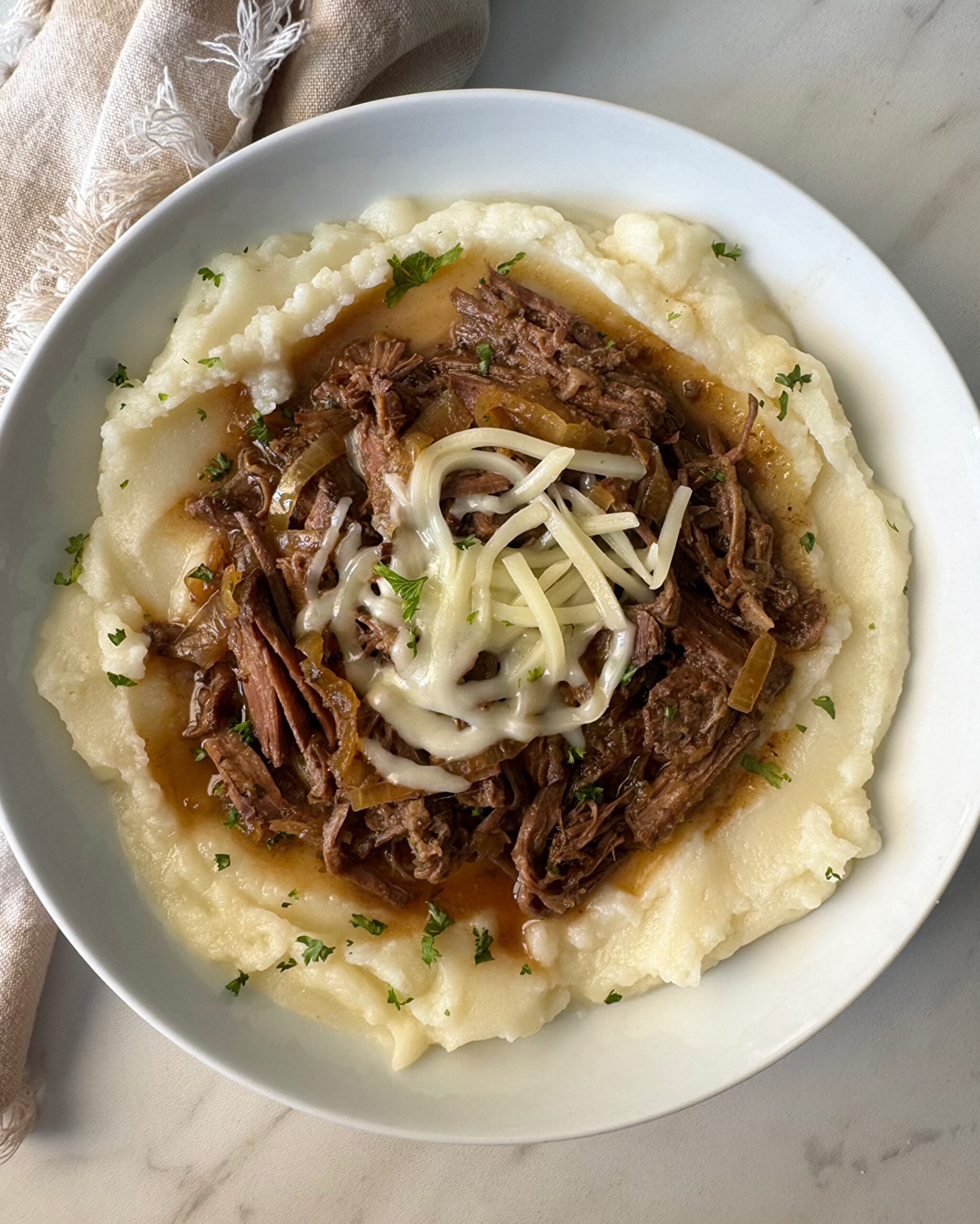 overhead shot of cooked French onion Pot Roast over mashed potatoes in a white bowl garnished with melted shredded cheese