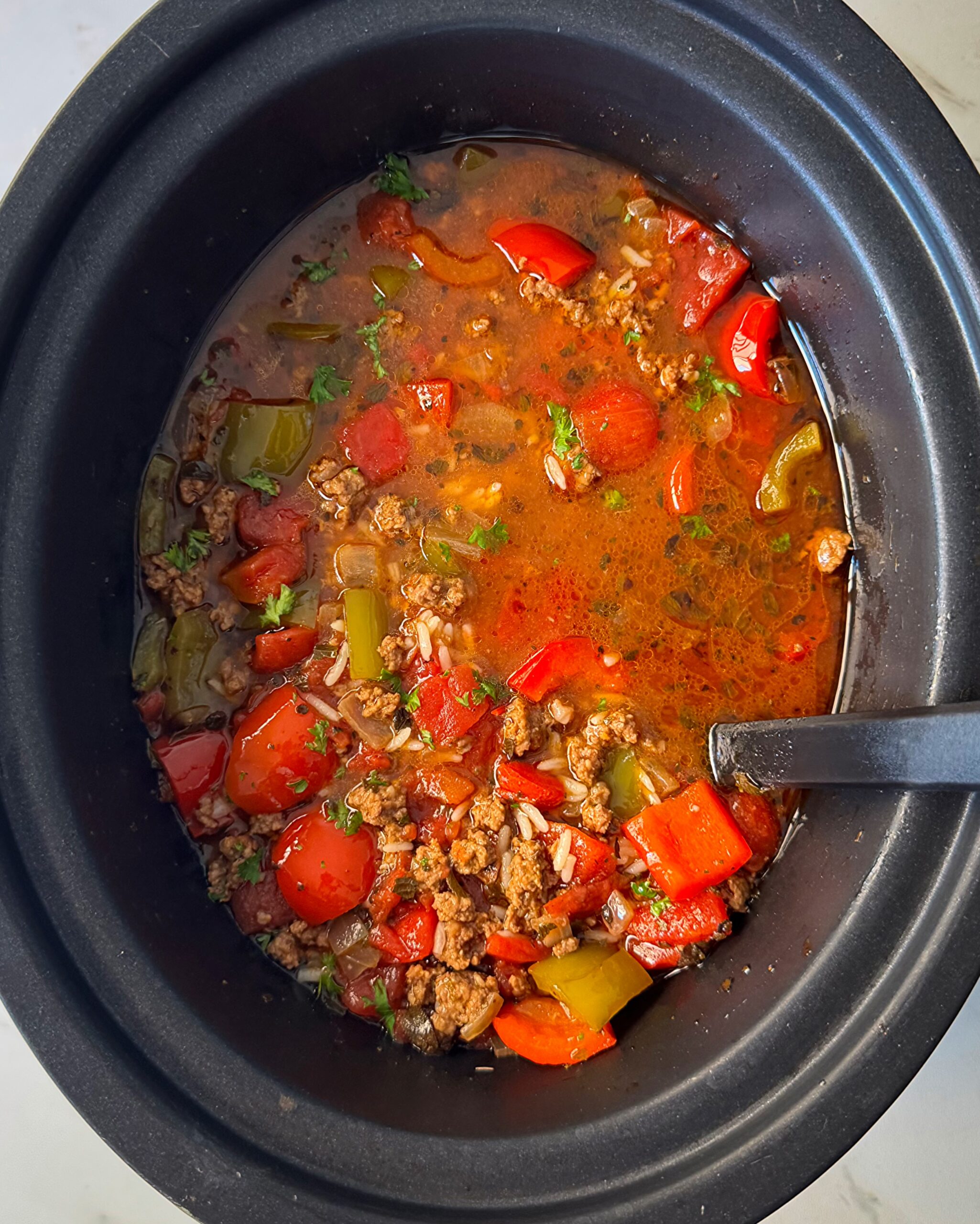 overhead shot of cooked Stuffed Pepper Soup in a black slow cooker with a black spoon in it
