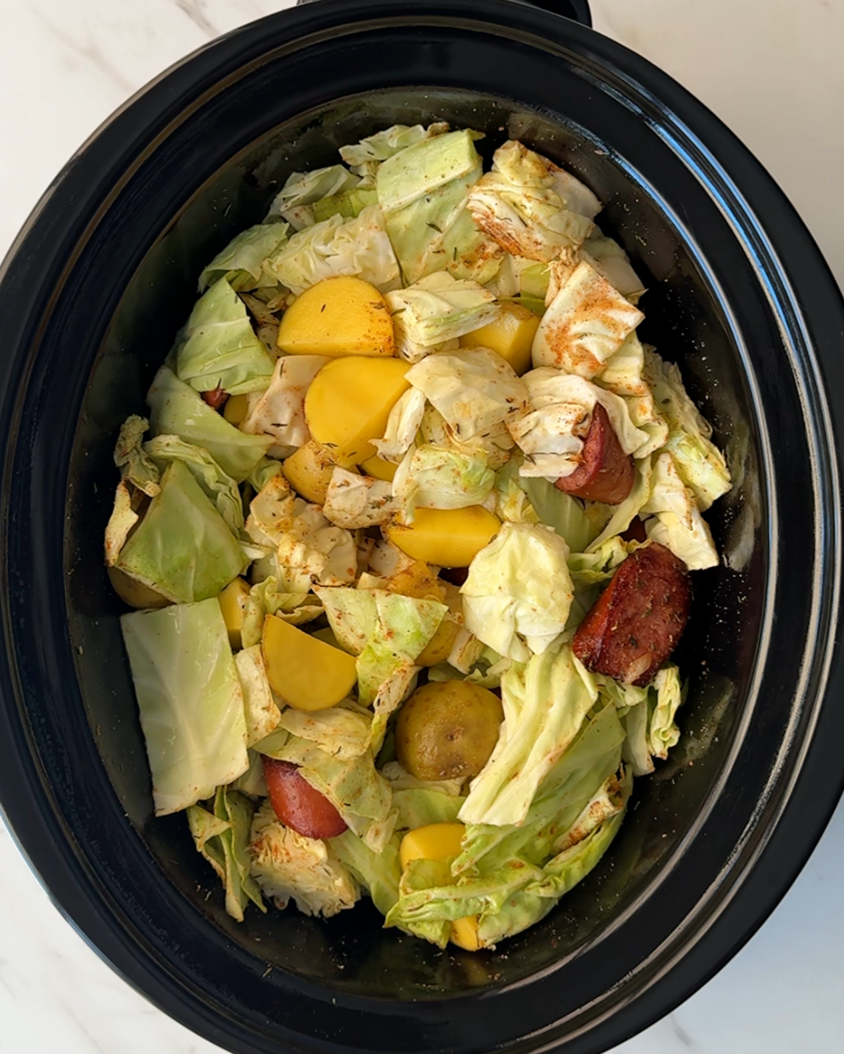 overhead shot of uncooked cabbage, sausage, and potatoes in a black slow cooker