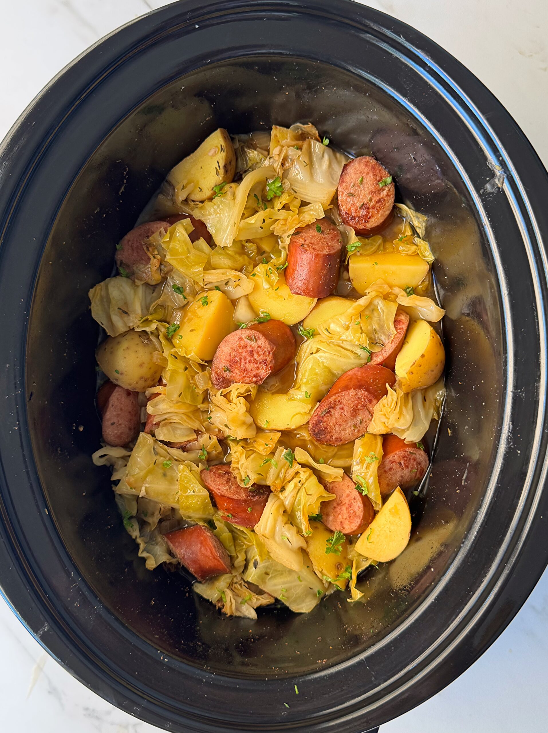 overhead shot of cooked cabbage, sausage, and potatoes in a black slow cooker