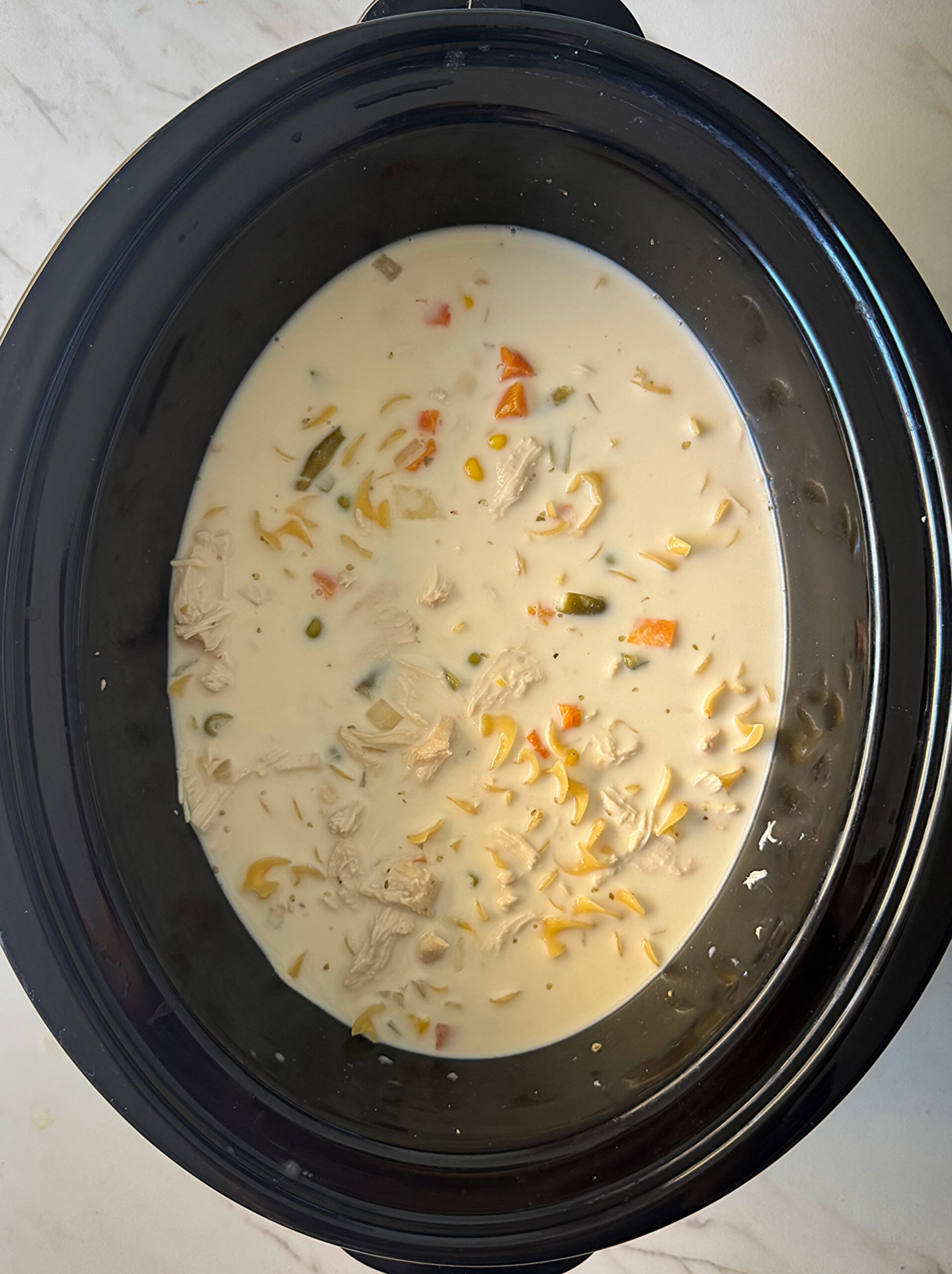 overhead shot of cooked chicken and noodles in a black slow cooker with cream and uncooked noodles