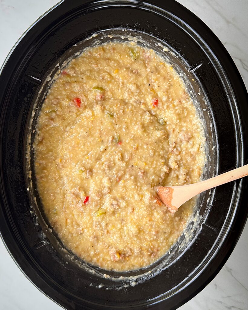 overhead shot of cooked sausage cheese grits in a black slow cooker with a wooden spoon