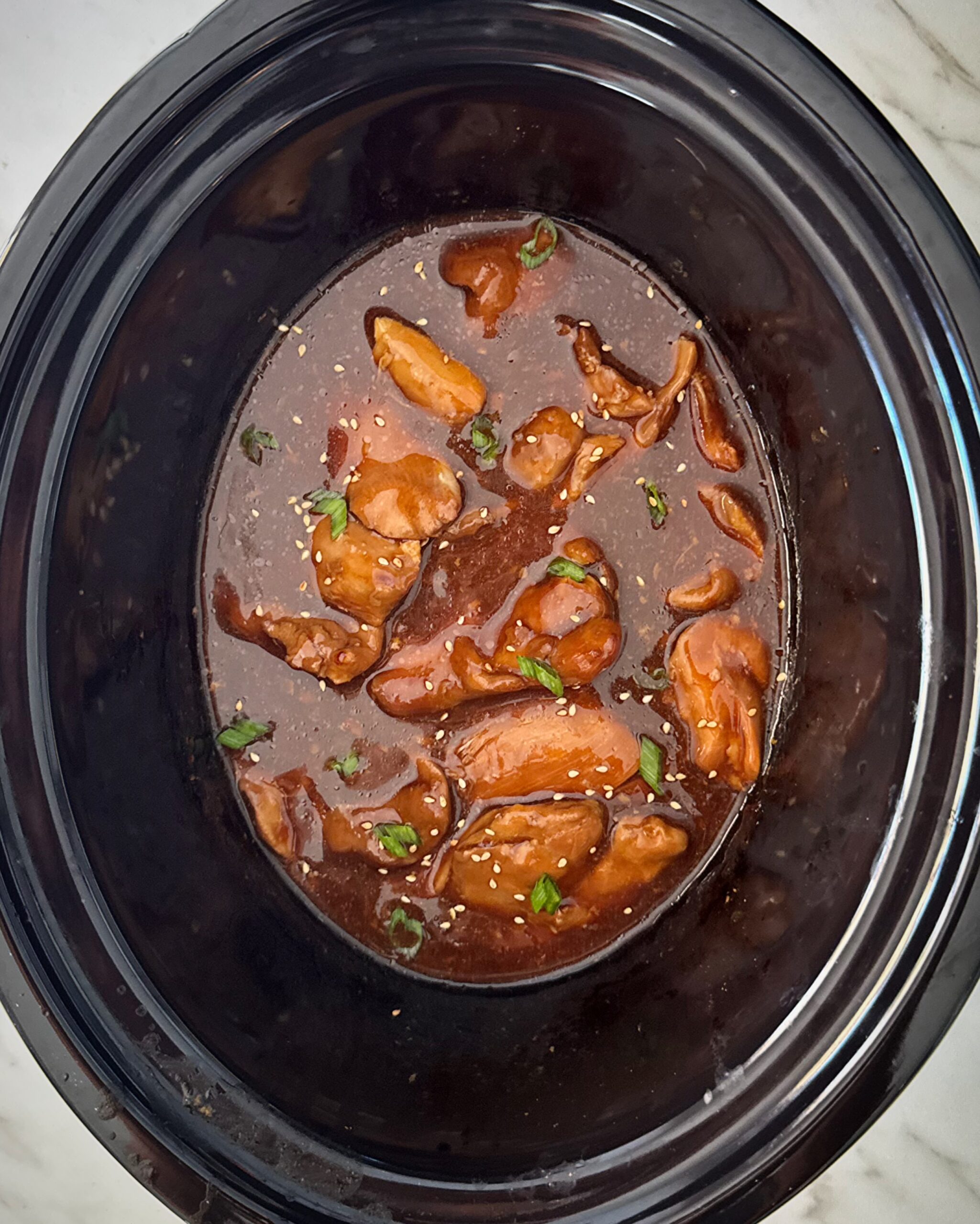 overhead shot of cooked teriyaki chicken in a black slow cooker garnished with green onion and sesame seeds