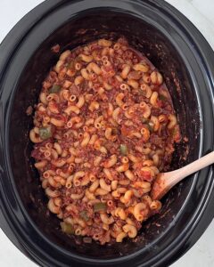 overhead shot of uncooked ingredients for American Goulash in a black slow cooker with pasta added