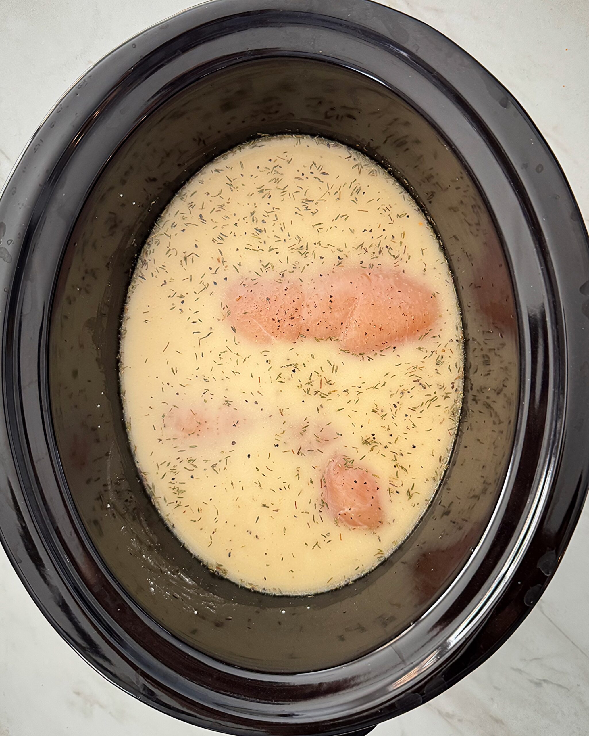 overhead shot of uncooked chicken and gravy in a black slow cooker