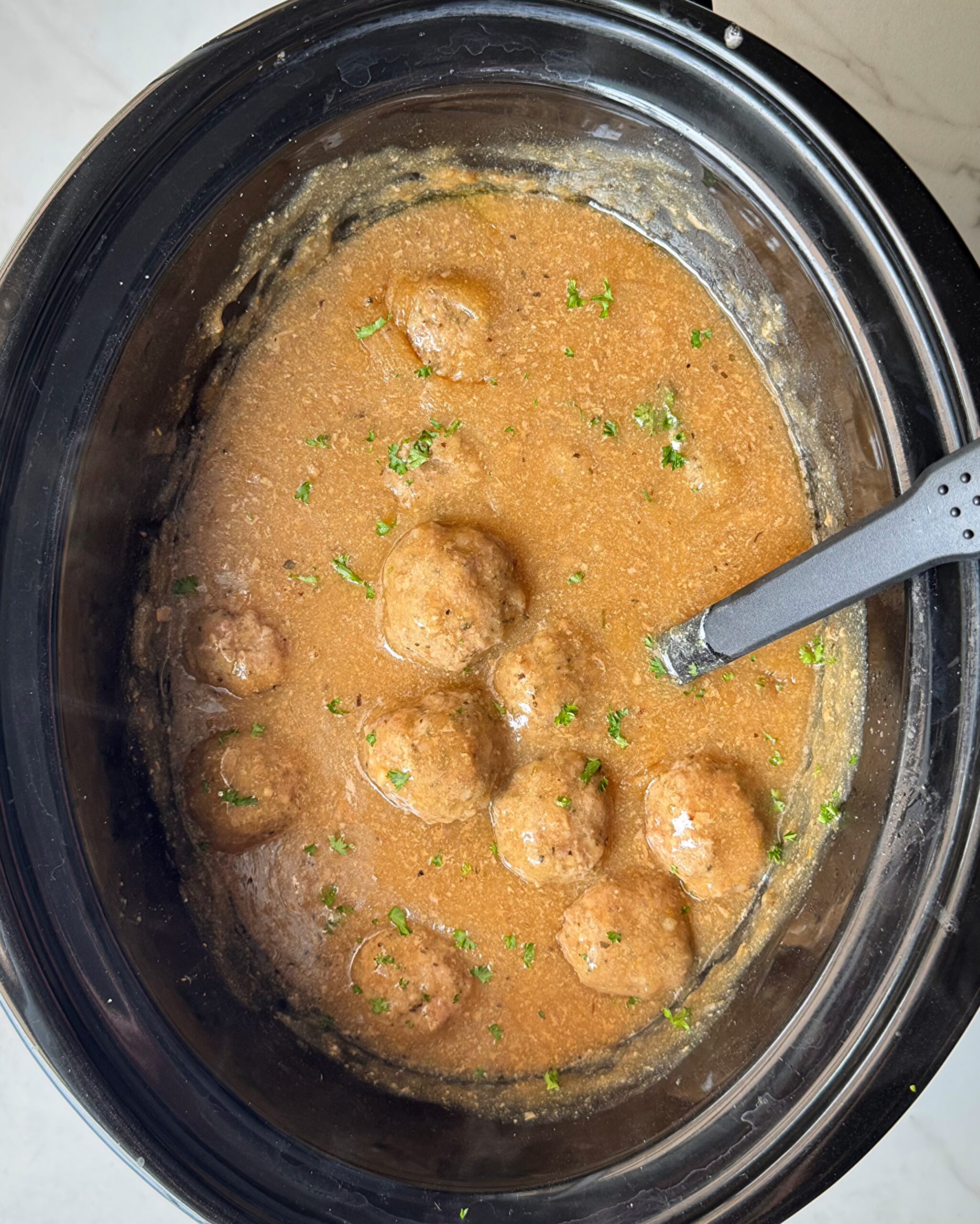 overhead shot of cooked meatballs and gravy in a black slow cooker garnished with fresh parsley