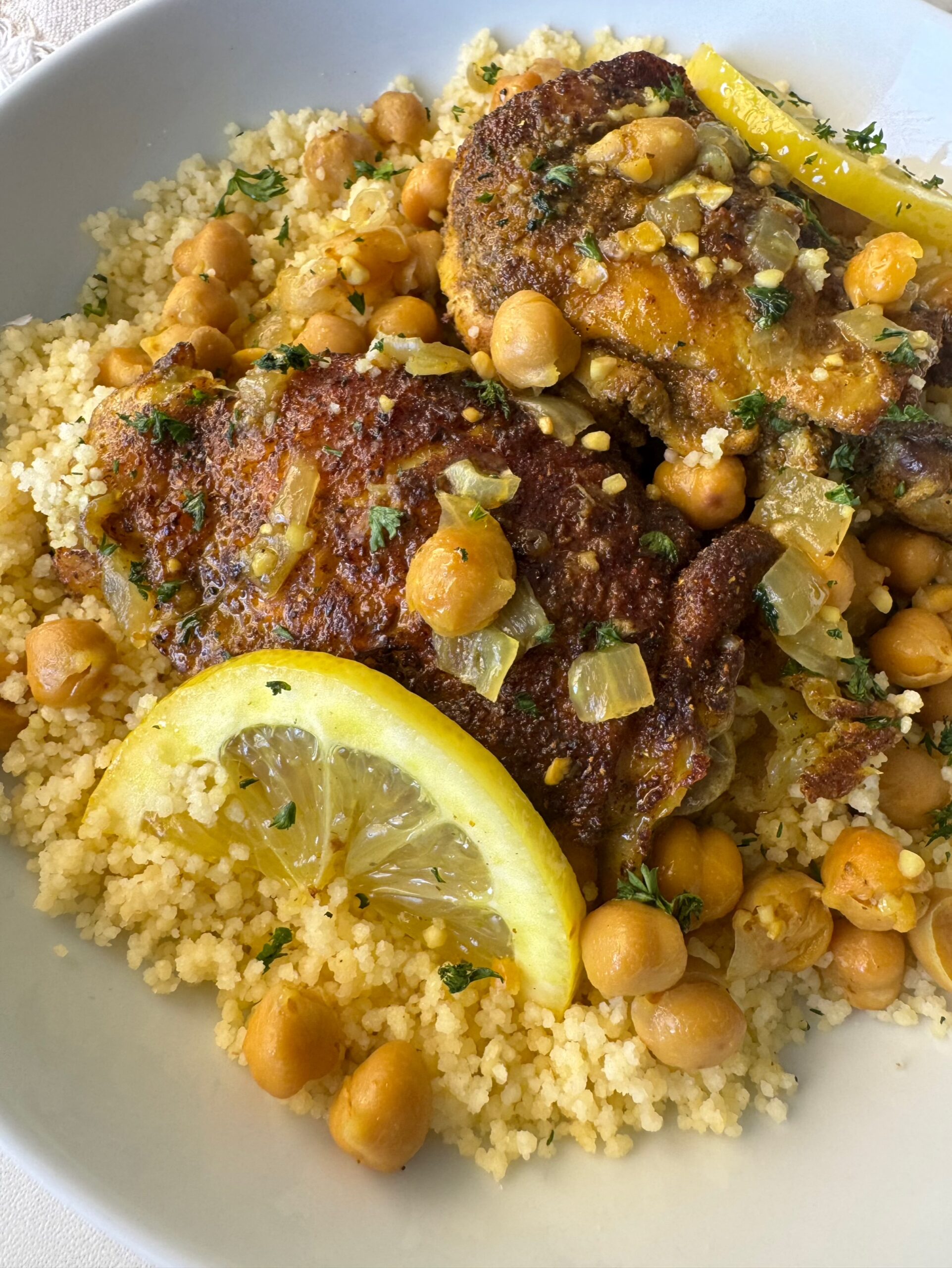 close up shot of cooked North African chicken on a white plate garnished with fresh parsley & sliced lemon served over couscous