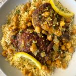 overhead shot of cooked North African chicken on a white plate garnished with fresh parsley & sliced lemon served over couscous