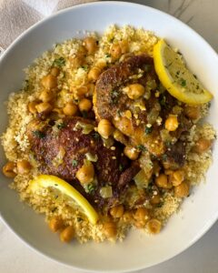 overhead shot of cooked North African chicken on a white plate garnished with fresh parsley & sliced lemon served over couscous