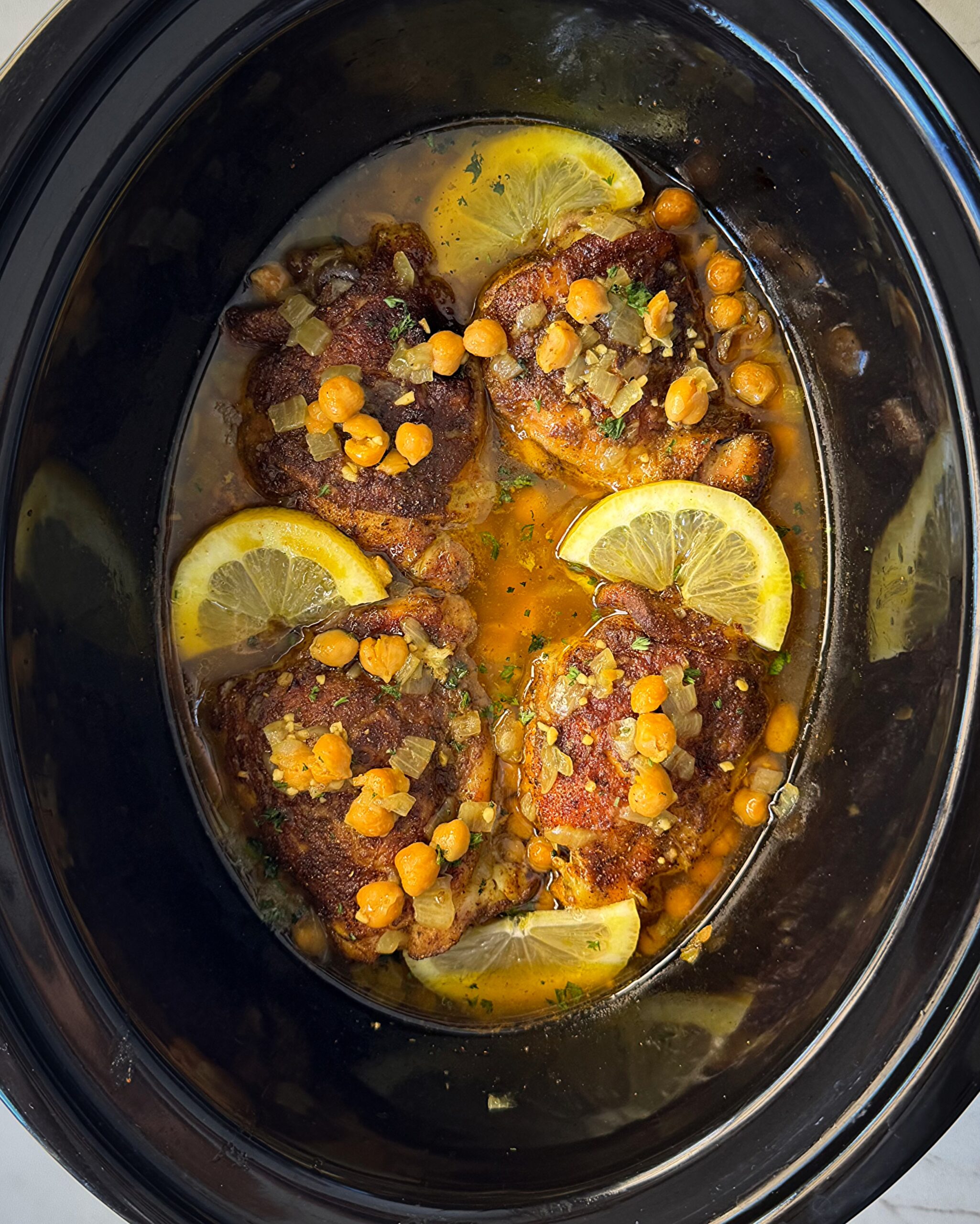 overhead shot of cooked North African chicken in a black slow cooker garnished with fresh parsley & sliced lemon