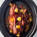overhead shot of cooked ham in a black slow cooker surrounded by diced pineapple