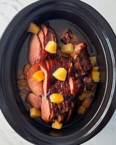 overhead shot of cooked ham in a black slow cooker surrounded by diced pineapple