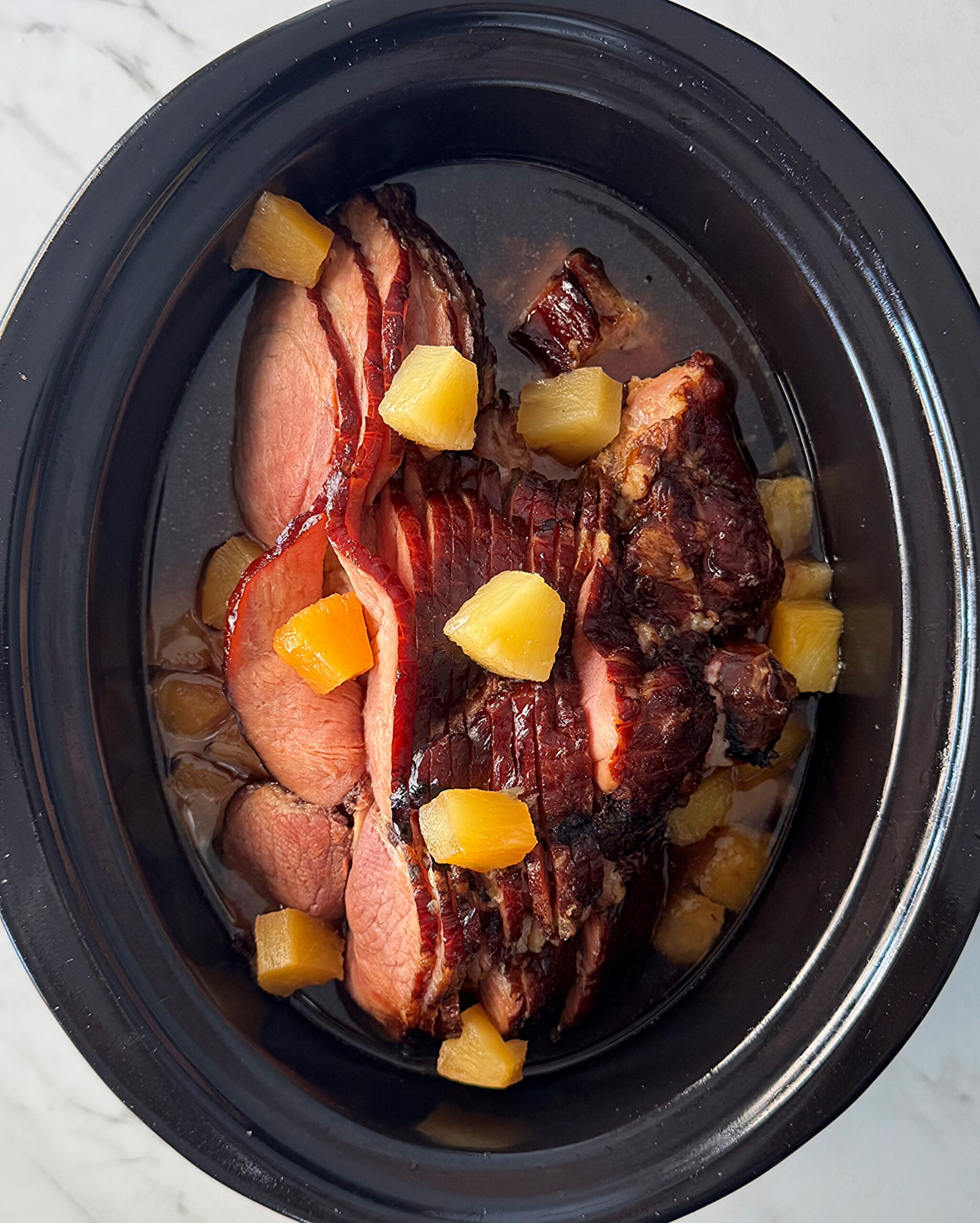 overhead shot of cooked ham in a black slow cooker surrounded by diced pineapple