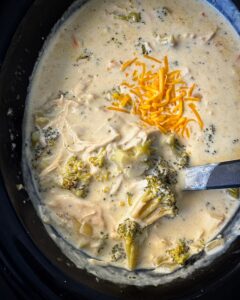 close up shot of cooked Chicken Broccoli Cheese Soup in a black slow cooker garnished with shredded cheddar cheese