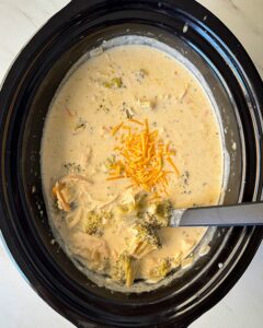 overhead shot of cooked Chicken Broccoli Cheese Soup in a black slow cooker garnished with shredded cheddar cheese