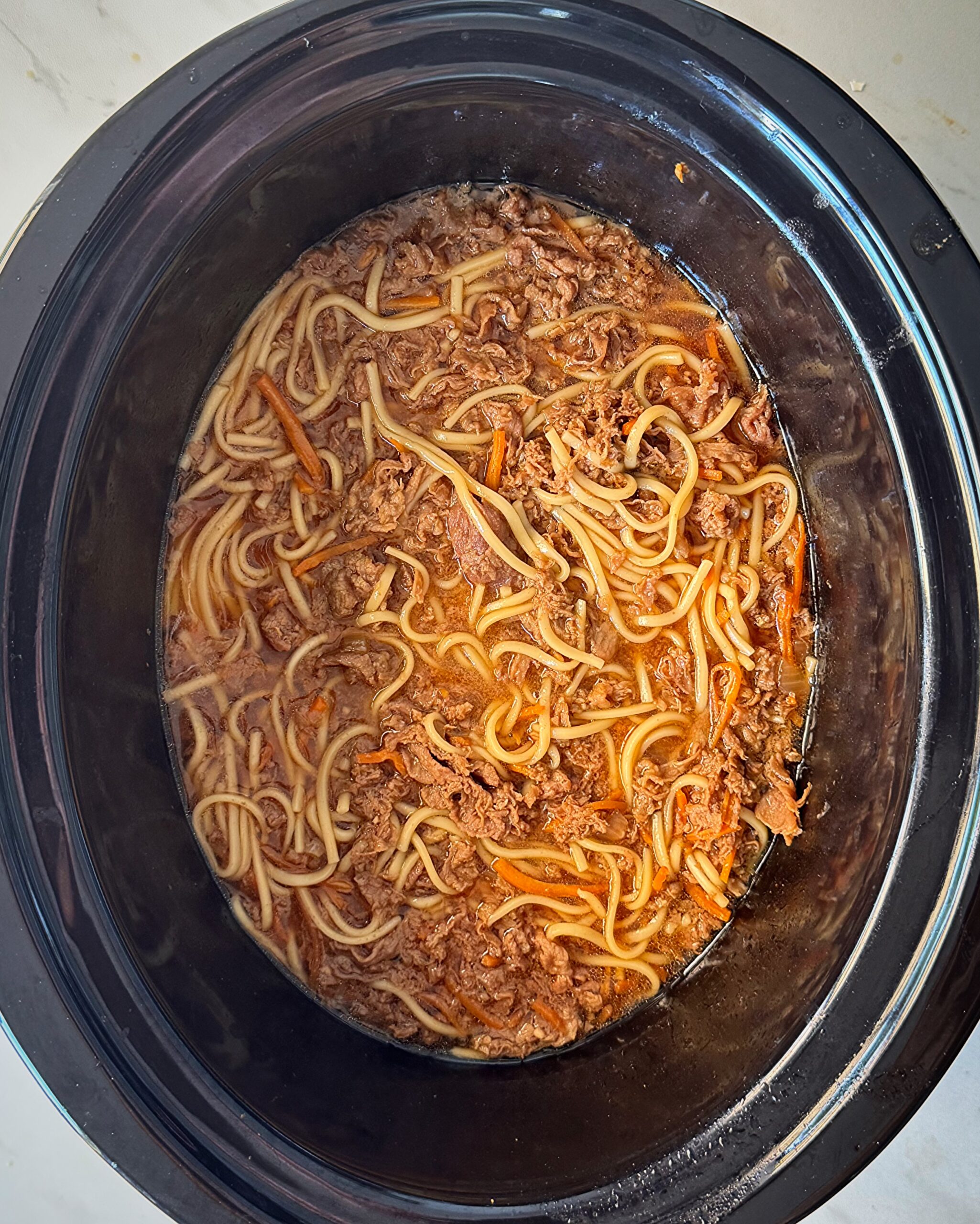 overhead shot of uncooked beef udon soup in a black slow cooker with noodles added