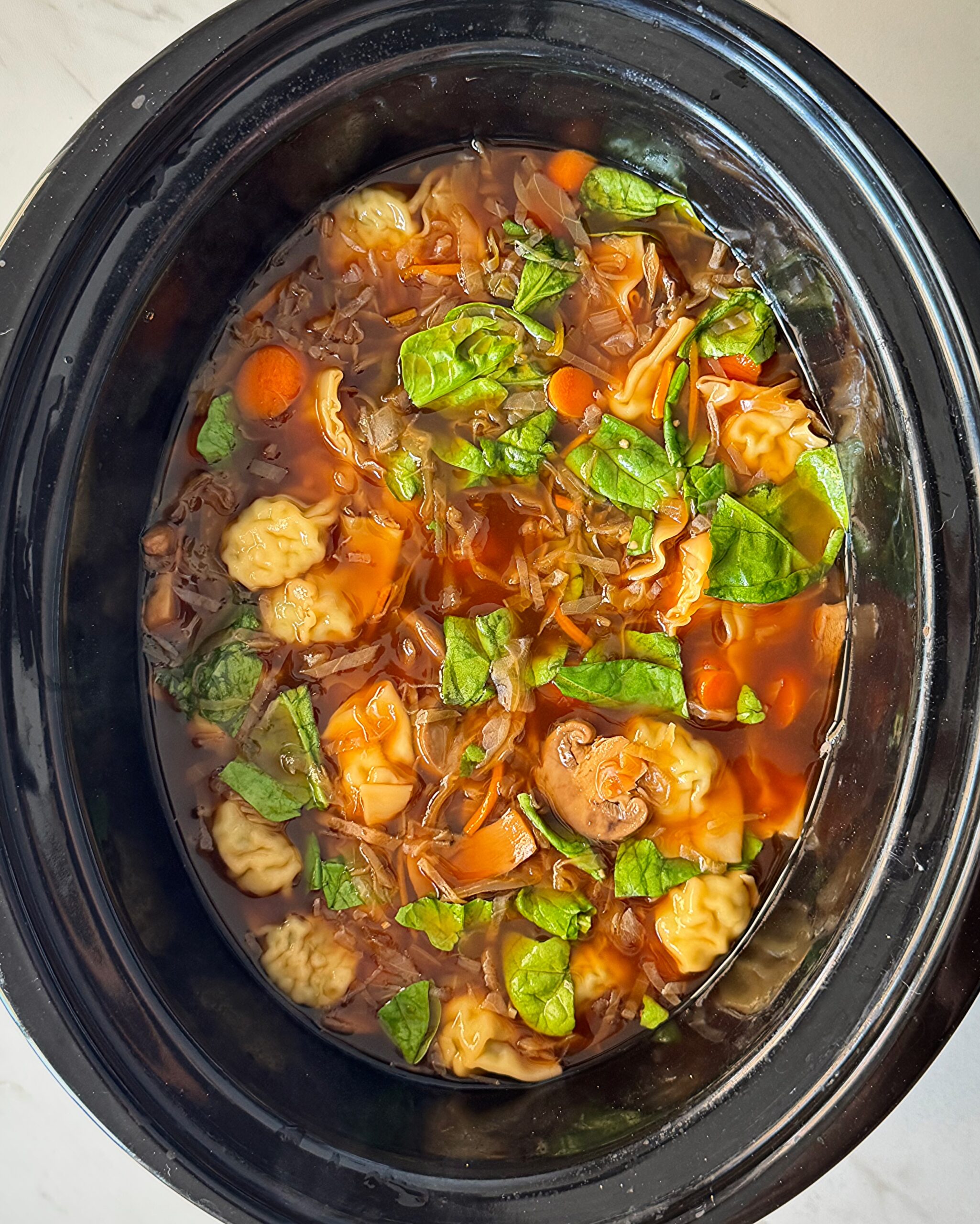 overhead shot of cooked potsticker soup in a black slow cooker with potstickers added and fresh spinach