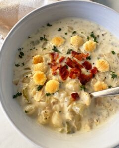 close up shot of cooked clam chowder in a white bowl garnished with fresh parsley, bacon, and oyster crackers